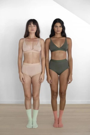 Two women standing side-by-side wearing matching bra and underwear sets in front of a white wall, full shot.