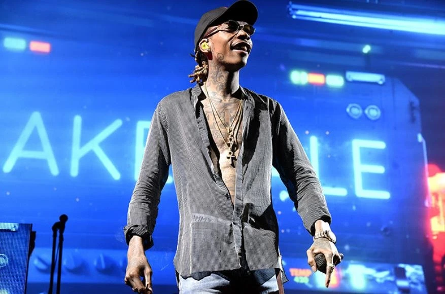 Wiz Khalifa performs on stage in an open black and gray vertical striped shirt, jeans, baseball cap, necklaces, and sunglasses.