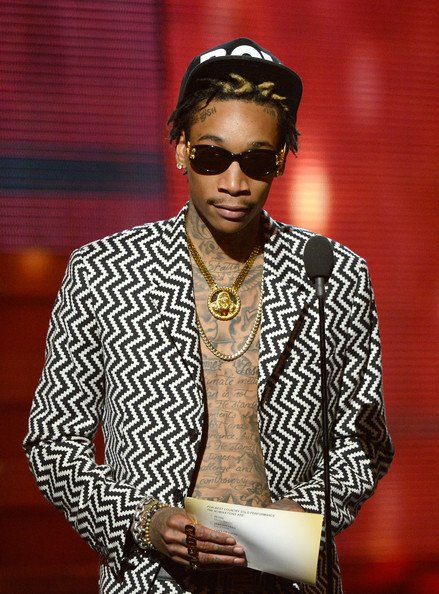 Wiz Khalifa on stage in a black-and-white patterned jacket, hat, and sunglasses, holding papers near a microphone at an event.