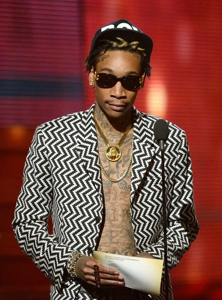 Wiz Khalifa on stage in a black-and-white patterned jacket, hat, and sunglasses, holding papers near a microphone at an event.