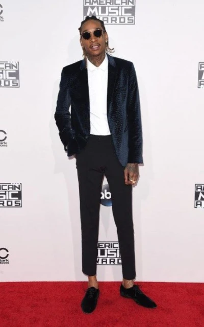 Adult man wearing a velvet blazer, dress pants, and sunglasses stands on a red carpet in front of a branded backdrop for an event.