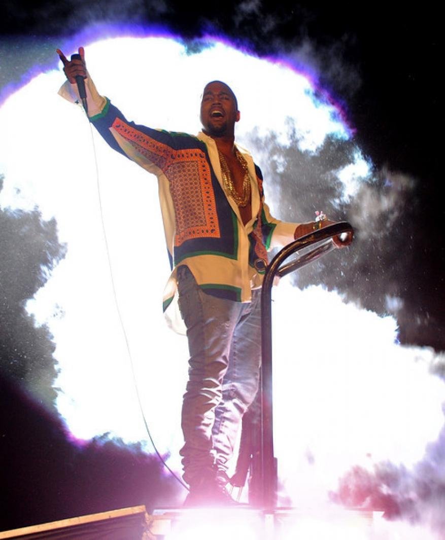 Male musician performing live on stage with a microphone, wearing an patterned shirt, jeans, and gold chain necklace, illuminated by stage lights.