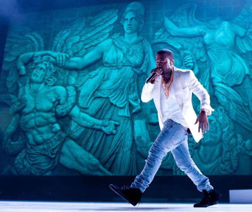 Man in white blazer sings into microphone on stage with large, turquoise bas-relief sculpture of Greek figures in background.