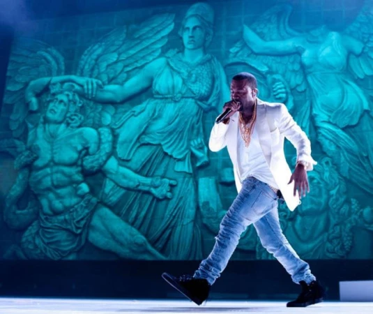 Man in white blazer sings into microphone on stage with large, turquoise bas-relief sculpture of Greek figures in background.