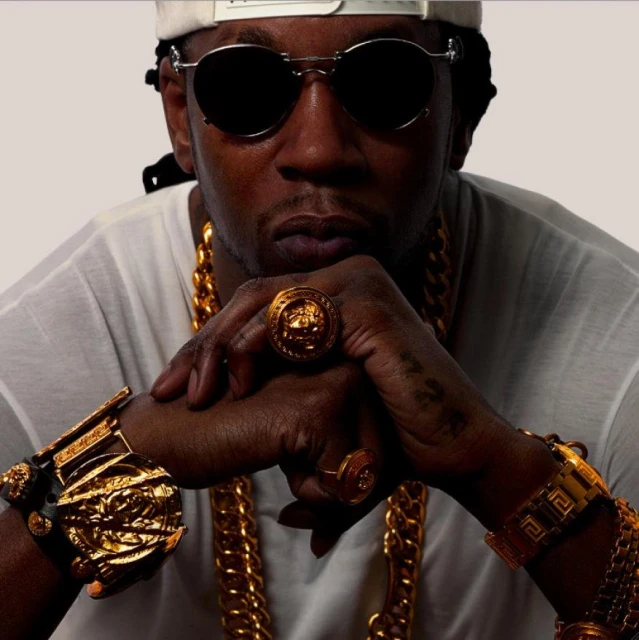 Portrait of an adult man wearing dark sunglasses and a white cap, adorned with gold jewelry, rings, and a large chain.