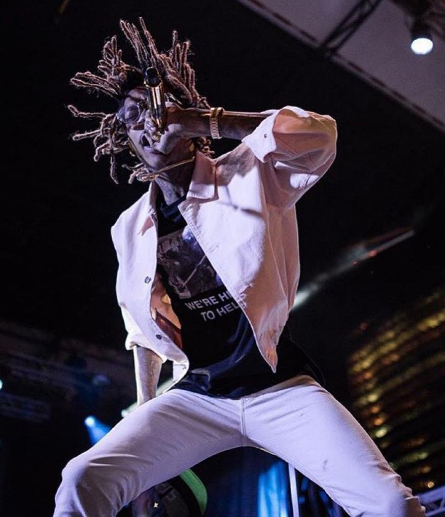 Rapper performing on stage, wearing a white jacket and jeans, holding a microphone, with a city building visible in background.