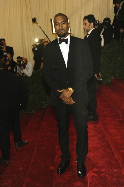 Man in a black tuxedo and bow tie standing on a red carpet. He has a gold watch on his wrist and is looking at the camera.