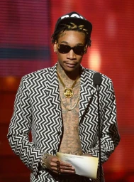 Wiz Khalifa wearing a black and white zigzag patterned blazer, dark sunglasses, and gold chains standing at a podium.