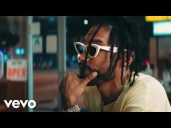 Man with dreadlocks wearing white sunglasses and yellow t-shirt seated inside at night with city lights visible through glass