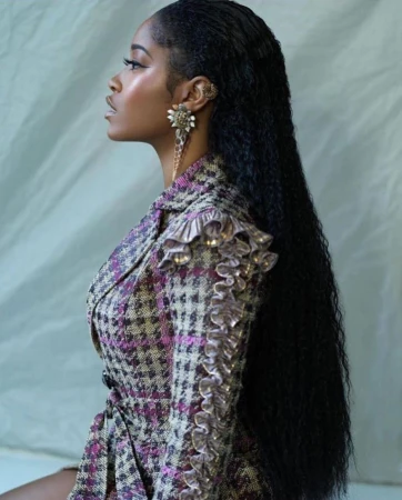 Profile portrait of a Black woman with long dark wet-look hair wearing a patterned blazer and statement floral earring against a draped backdrop