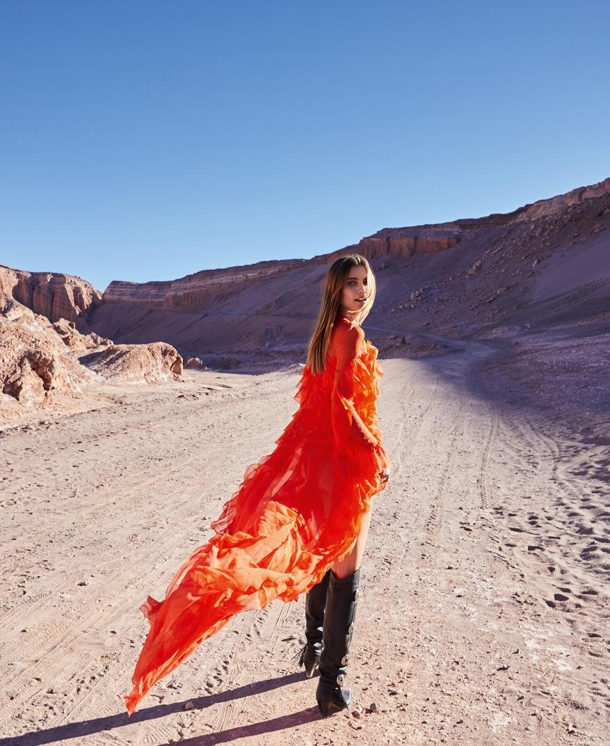 Woman in an orange dress and black boots in a desert landscape under a blue sky. Fashion shot with natural light and minimal styling.