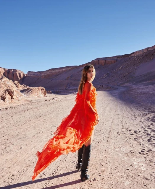 Woman in an orange dress and black boots in a desert landscape under a blue sky. Fashion shot with natural light and minimal styling.