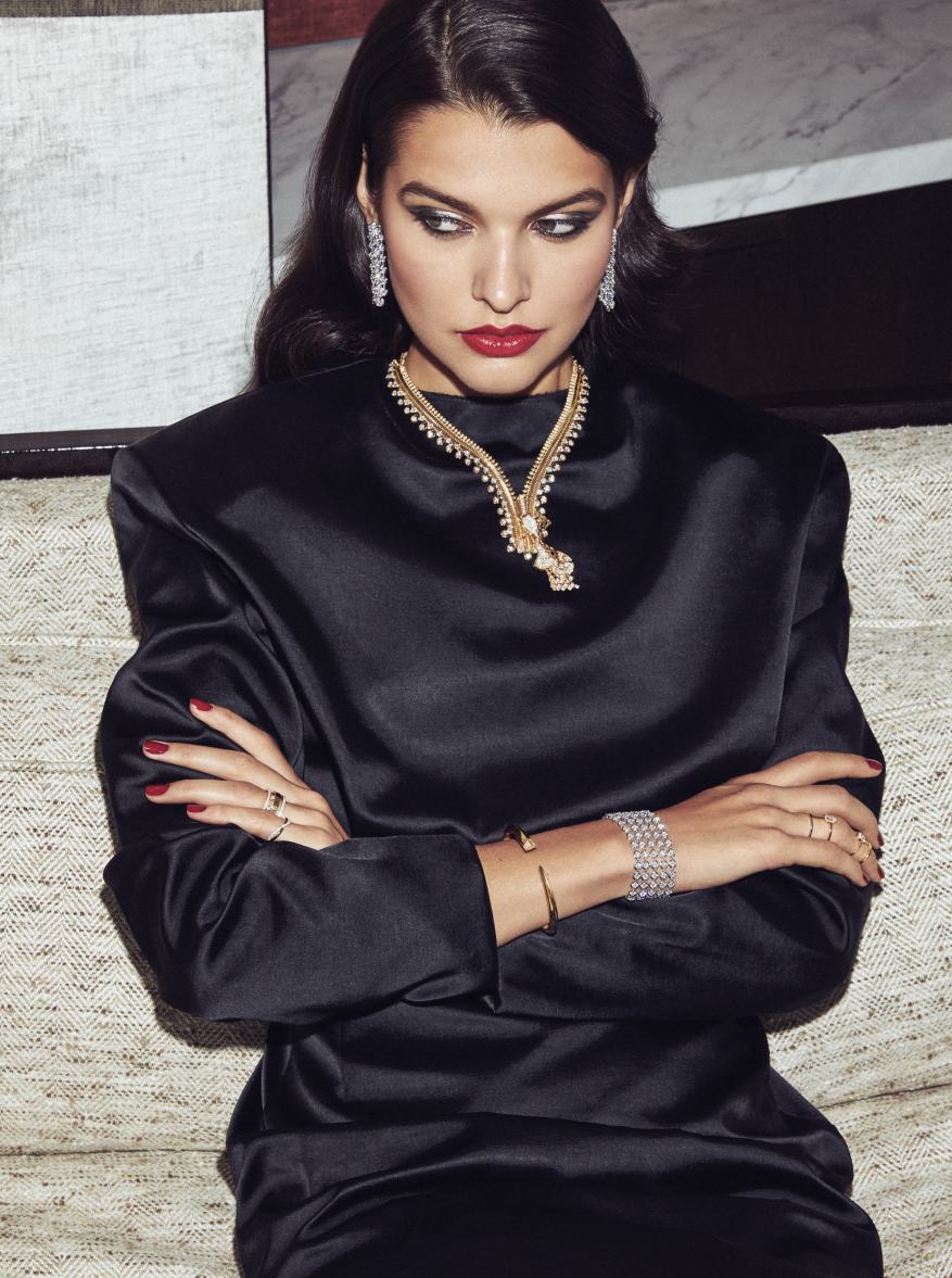 Brunette woman with red lipstick poses with diamond earrings, necklace and bracelets wearing black silk dress in indoor setting.