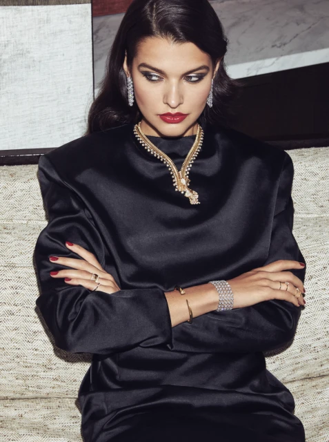 Brunette woman with red lipstick poses with diamond earrings, necklace and bracelets wearing black silk dress in indoor setting.