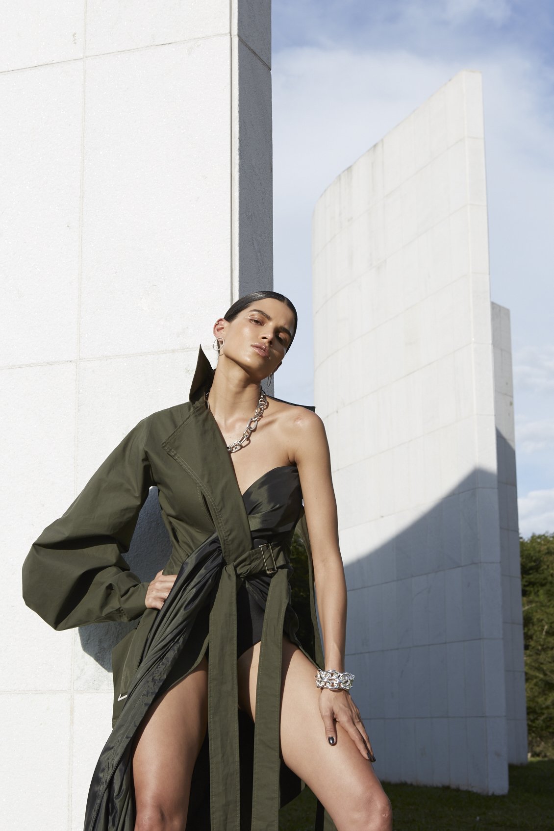 A woman poses in a dramatic high-slit olive dress with a black bodice in front of white, geometric architecture under a blue sky.