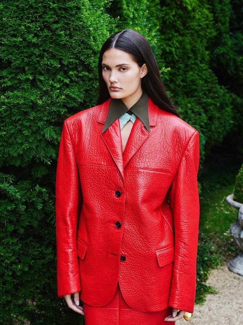 An adult female poses outdoors wearing a red blazer and skirt with a green bush background in a fashion editorial shot.
