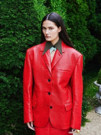 An adult female poses outdoors wearing a red blazer and skirt with a green bush background in a fashion editorial shot.