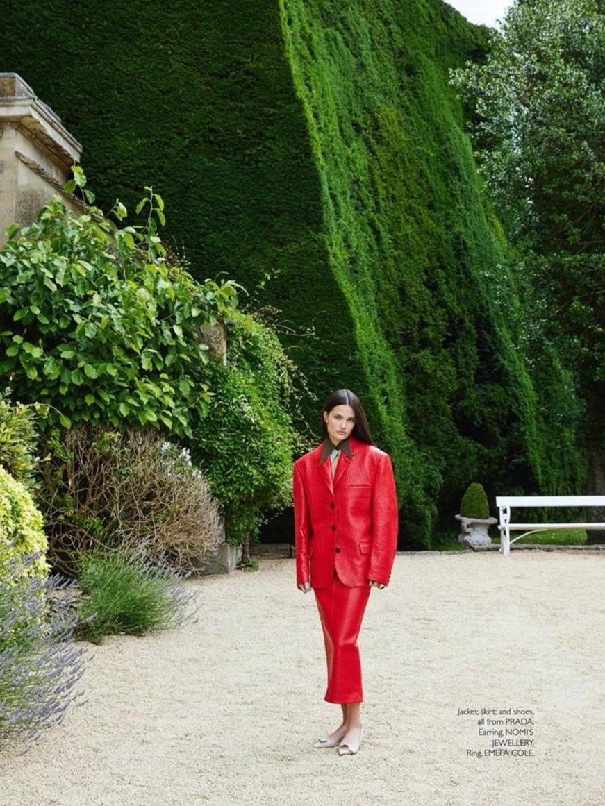 Fashion editorial shot outdoors features an adult female model in a red blazer and pants standing in a landscaped garden setting.