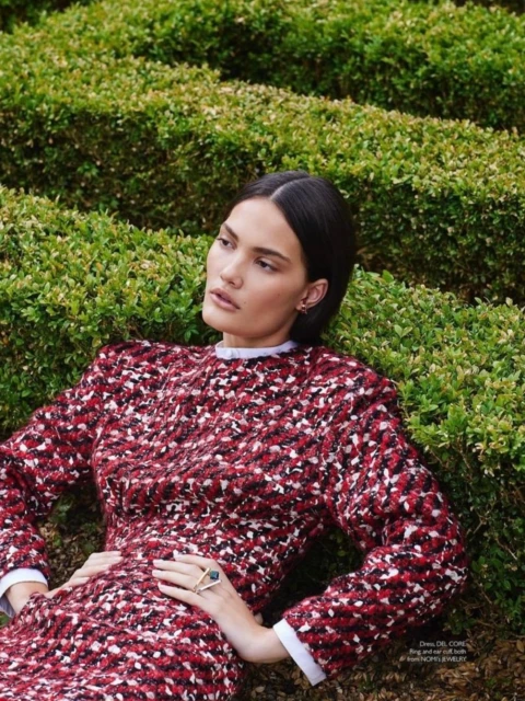 A woman poses in a red and white dress, lying amongst green bushes. A large ring adorns her finger, set against a backdrop of verdant foliage.
