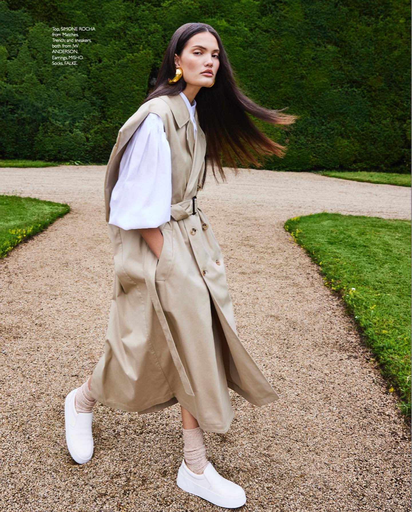 Editorial fashion shot: a woman in a trench coat walks on a gravel path with green foliage in the background, showcasing golden earrings.