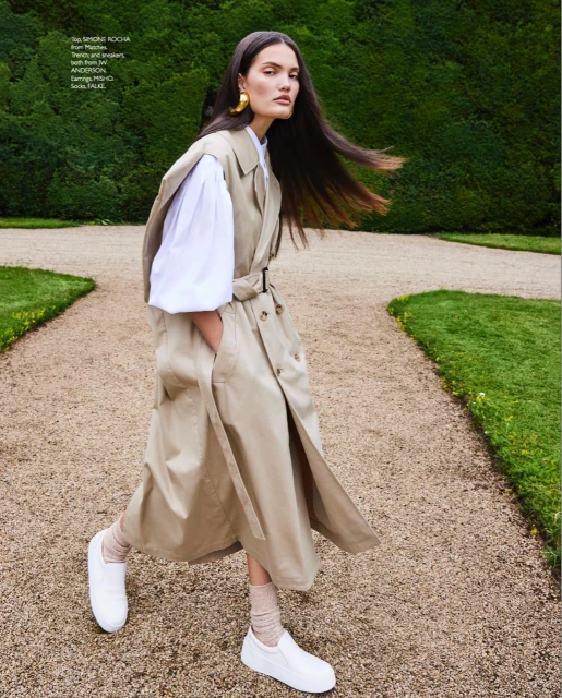 Editorial fashion shot: a woman in a trench coat walks on a gravel path with green foliage in the background, showcasing golden earrings.