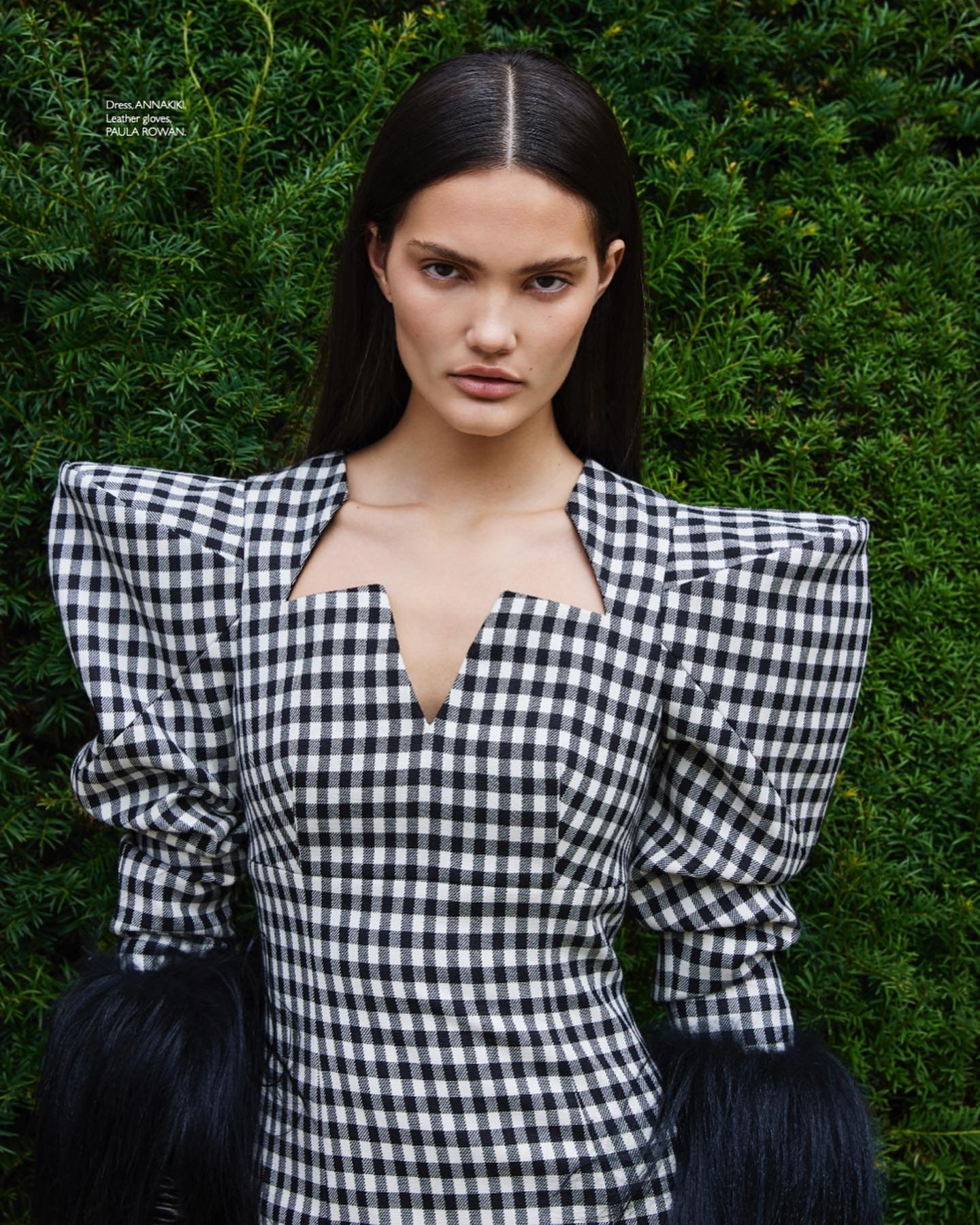 Fashion model wears a black and white checkered dress with fur sleeves, posing in front of a green hedge background.