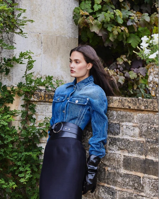 A woman in a denim jacket and black skirt poses against a stone wall, styled with wide belt and gloves, in an outdoor setting.