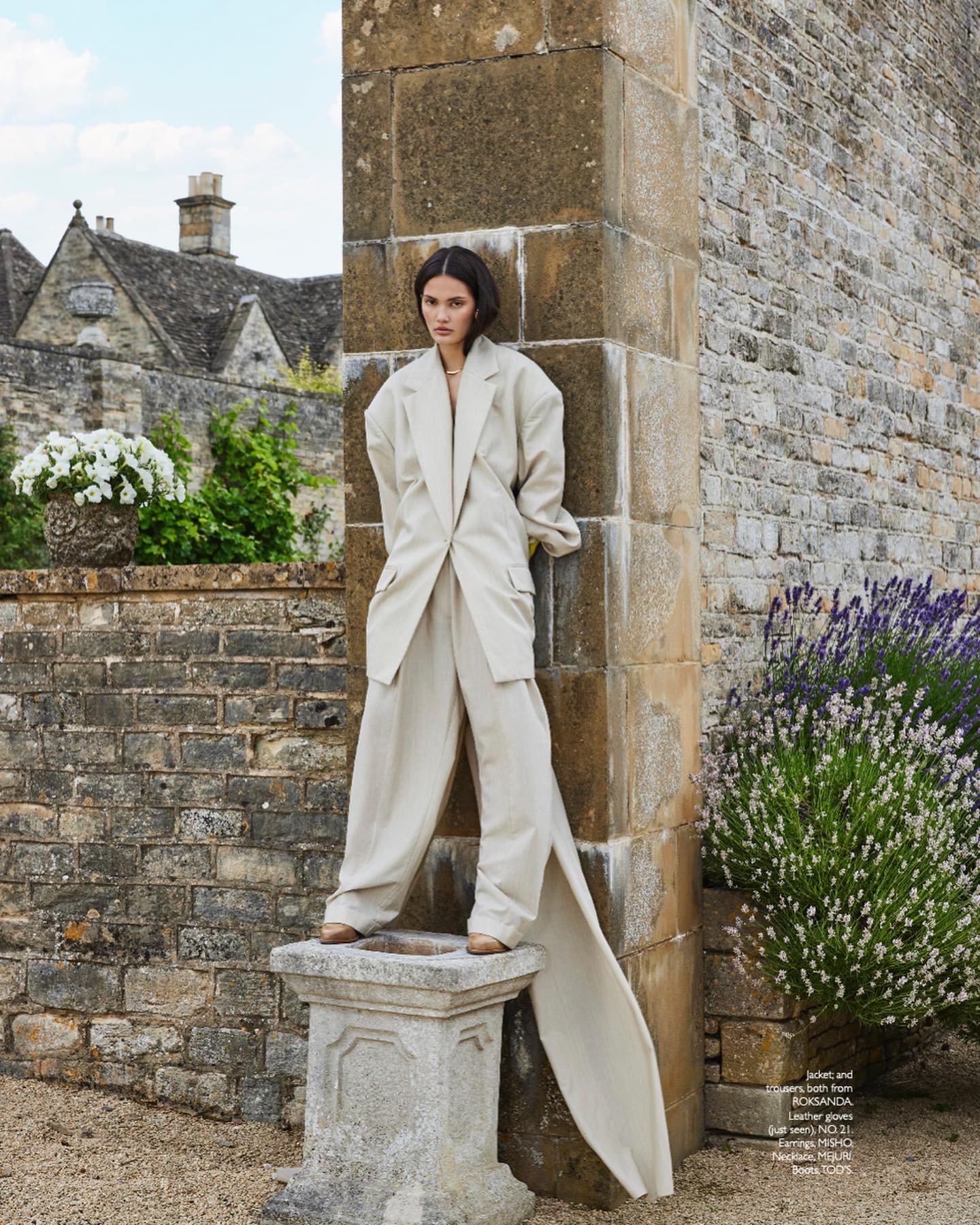 Editorial fashion shot of a woman in a beige suit standing on a stone plinth with a stone building and lavender in the background.