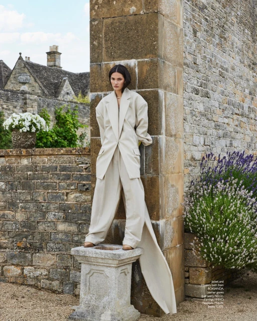 Editorial fashion shot of a woman in a beige suit standing on a stone plinth with a stone building and lavender in the background.