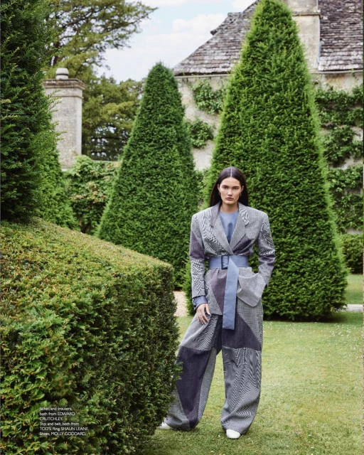 Fashion editorial shot of a female model in a patterned blazer and wide-leg pants, posing in a manicured garden with greenery and a building.