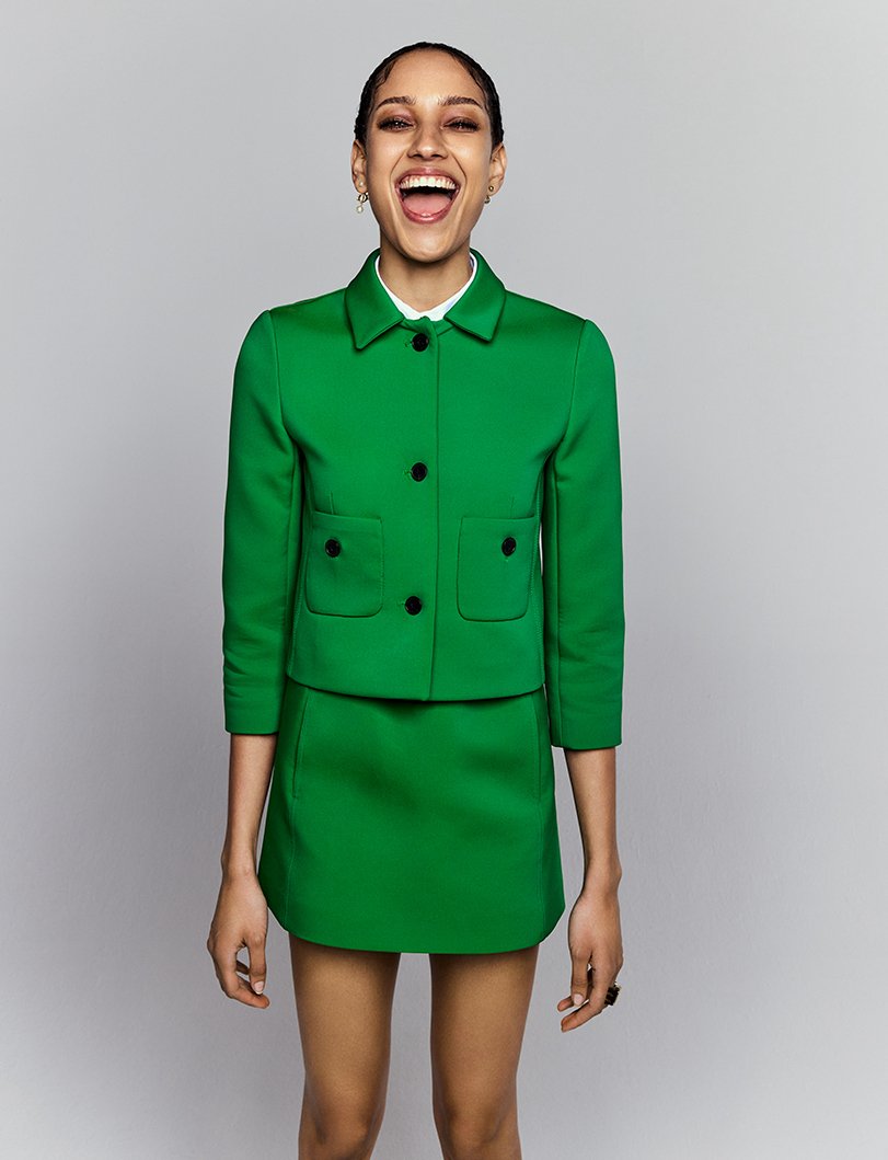 Woman smiles broadly in a bright green skirt suit against a gray background, showcasing a cheerful and fashionable studio portrait.
