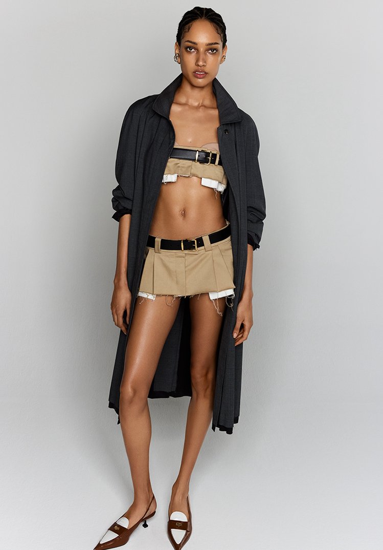 Fashion shot of a slender woman with tan skin, wearing a short skirt, top, jacket, and shoes, posing in a photo studio.