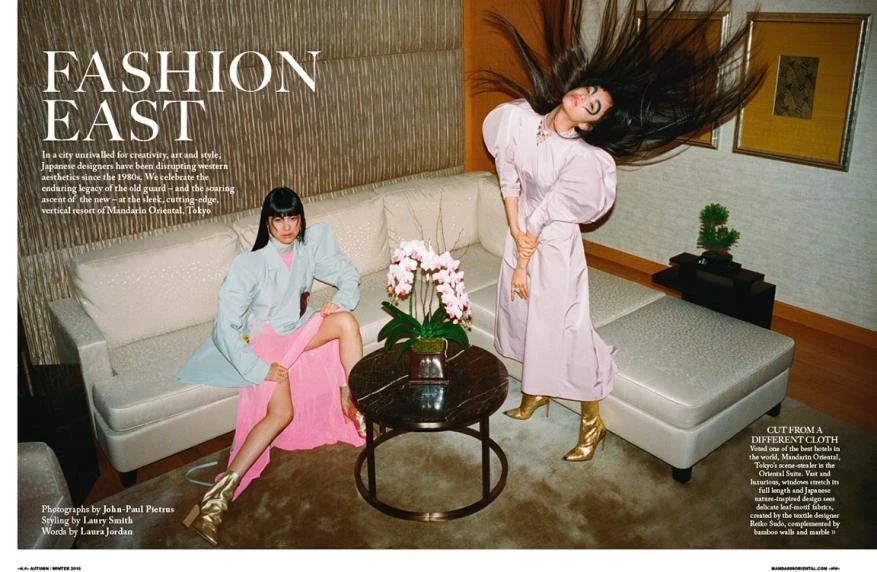 Two women pose in an interior setting for a fashion editorial, showcasing modern Japanese designers, with soft lighting and neutral tones.