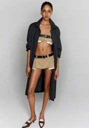 Full-length fashion portrait of a female model wearing a long dark coat, cropped top, pleated mini skirt, and flat shoes against a light grey background.
