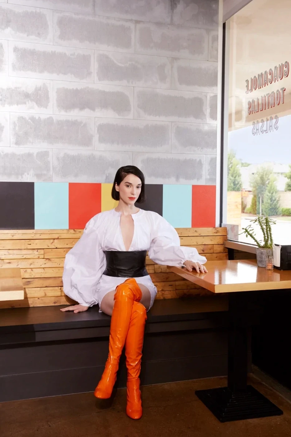 Woman with short dark hair in a white blouse and orange boots sitting at a wooden booth styled by Laury Smith
