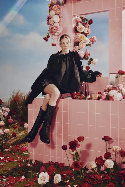 Fashion shot of a woman in a black dress and boots sitting on a pink tiled ledge with scattered roses, under a blue sky.
