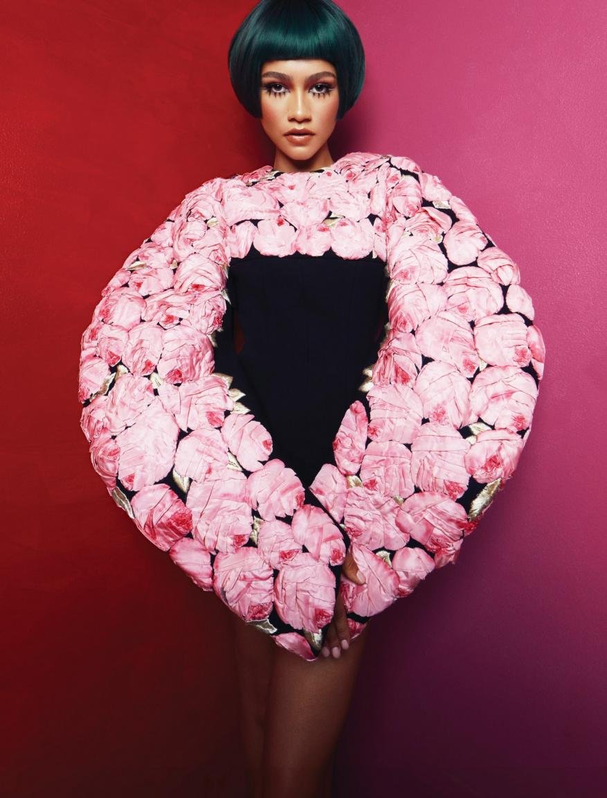 Zendaya wearing a couture rose-covered top with green hair and posing against a vibrant red and pink background in a studio setting.