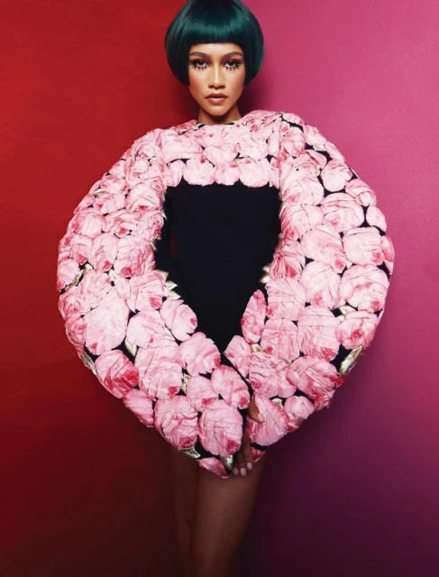 Zendaya wearing a couture rose-covered top with green hair and posing against a vibrant red and pink background in a studio setting.