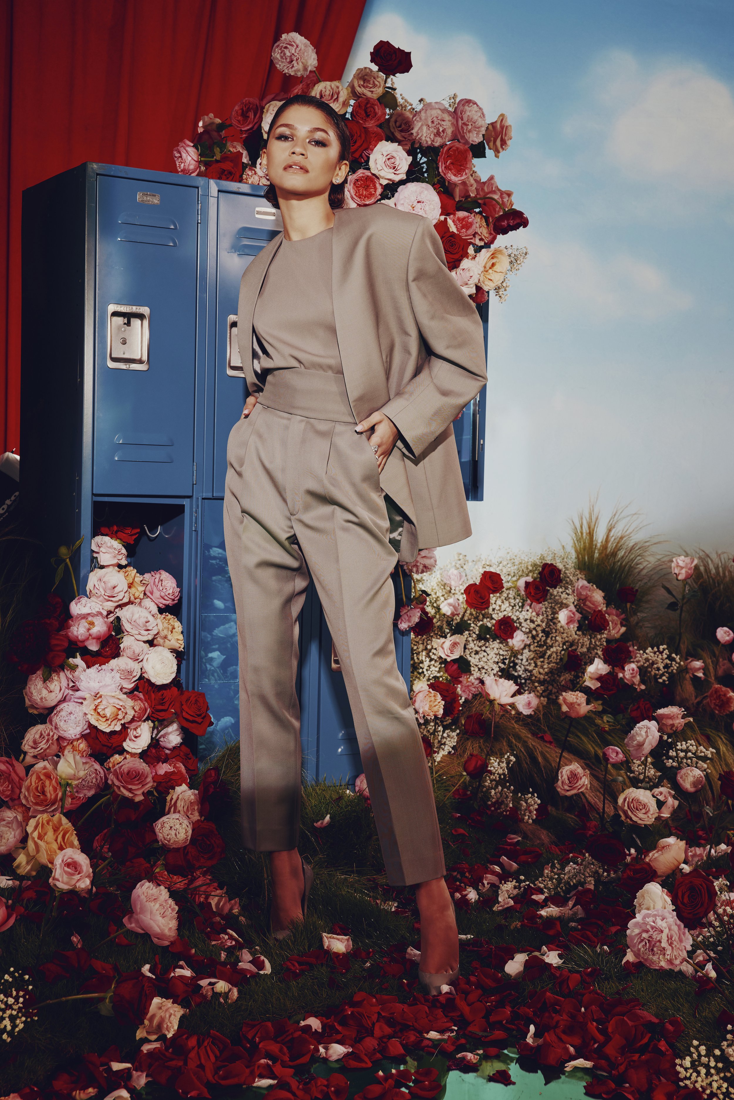 Zendaya poses in a tan suit amidst a floral arrangement with red curtains and blue lockers in the background, creating a surreal fashion portrait.