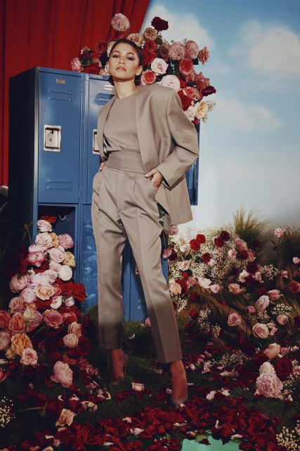 Zendaya poses in a tan suit amidst a floral arrangement with red curtains and blue lockers in the background, creating a surreal fashion portrait.