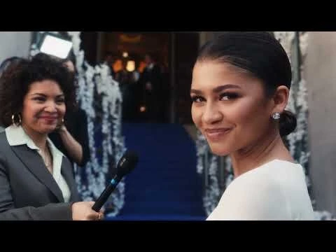 Zendaya smiles while being interviewed on the red carpet, posing in a white dress alongside a reporter holding a microphone.