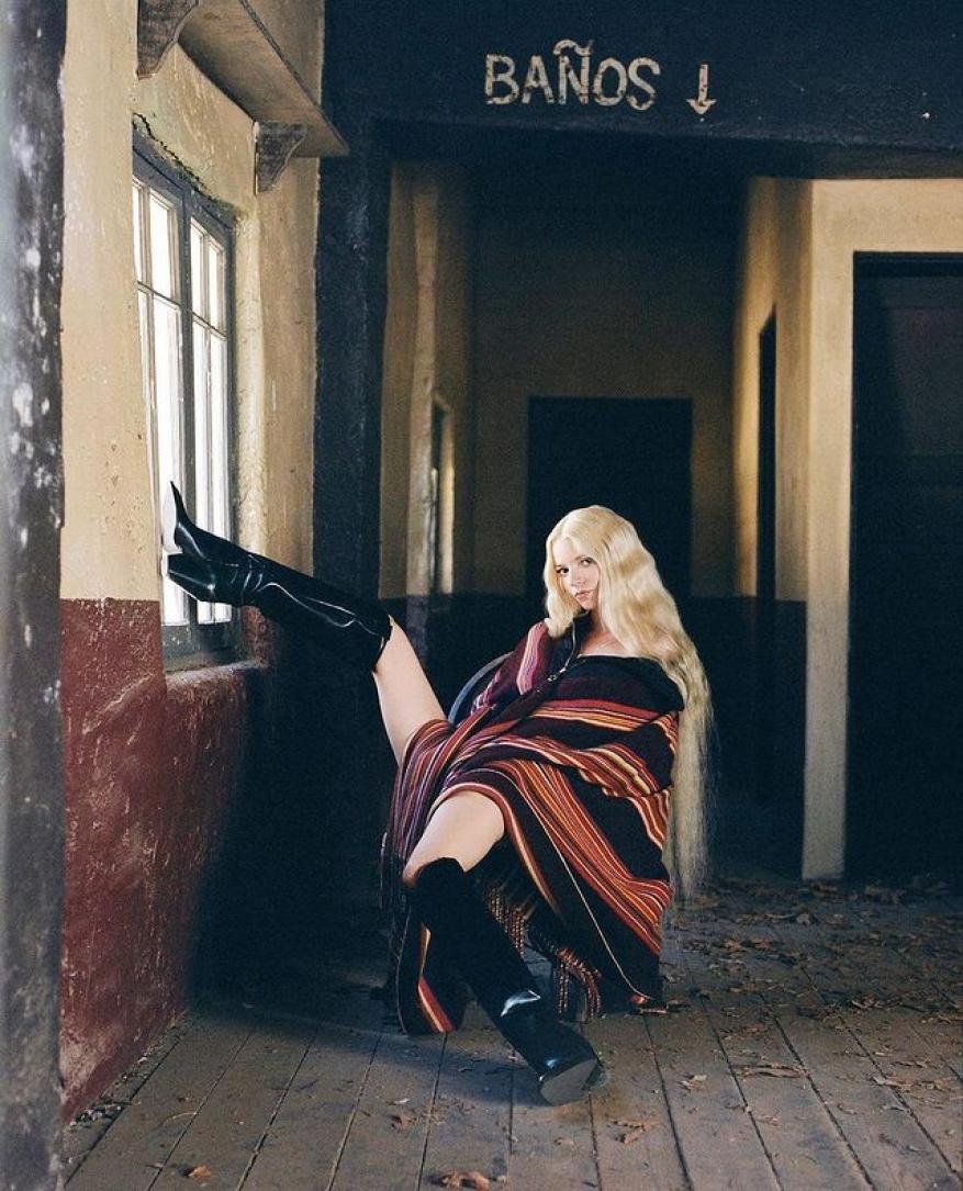 Blonde woman in high-shine boots poses near a window in a rustic building, draped in a colourful, striped blanket.
