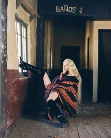 Blonde woman in high-shine boots poses near a window in a rustic building, draped in a colourful, striped blanket.