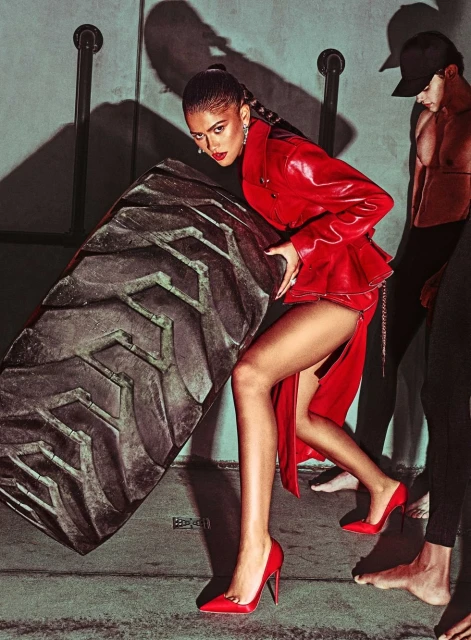 Woman in red dress and heels pushes large tire in industrial setting with other people standing in the background.