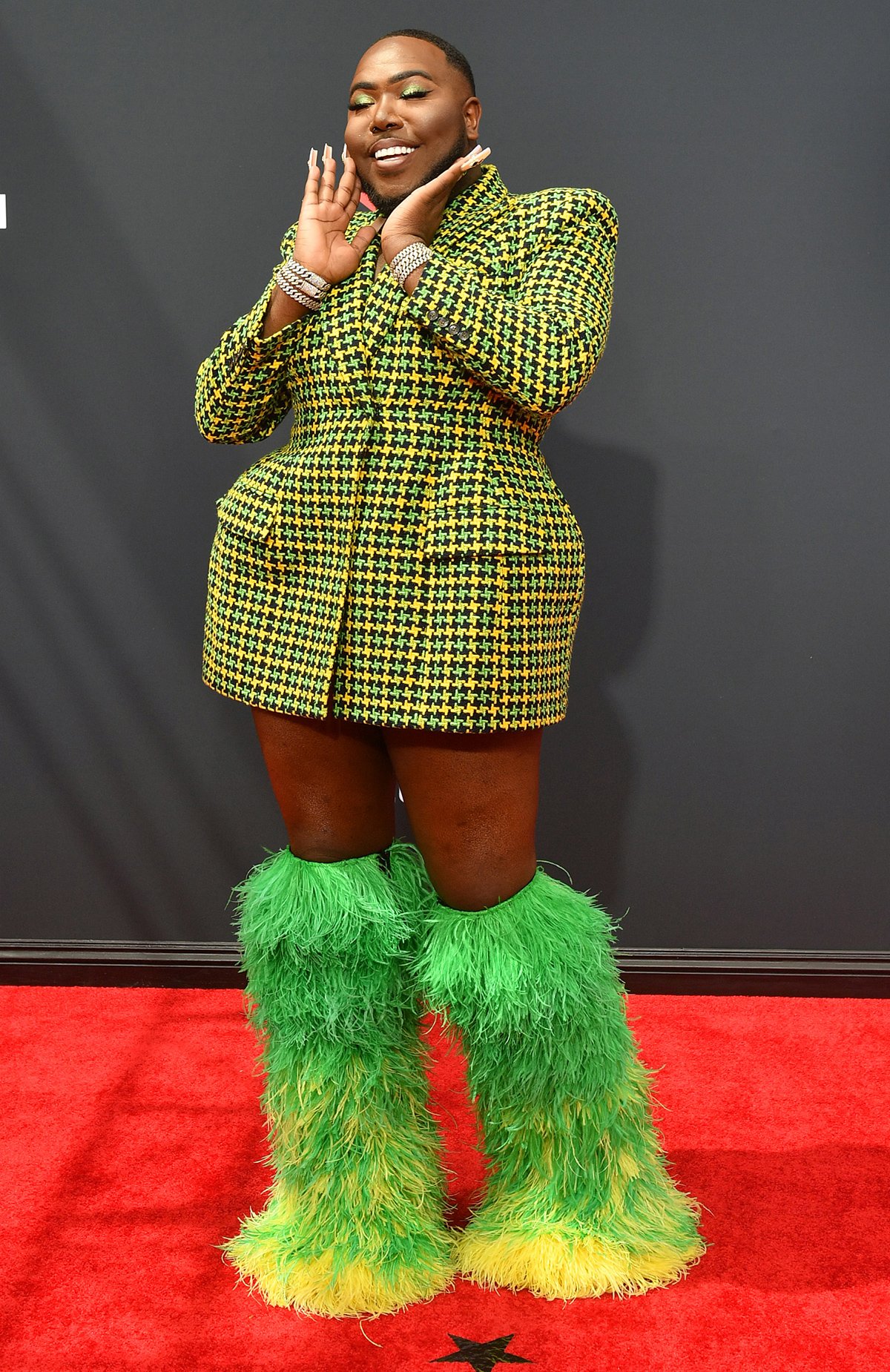 A person poses on a red carpet in a yellow and green checkered blazer dress and large furry green leg warmers.