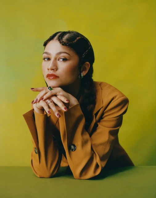 Portrait of Zendaya wearing a brown suit with green rings and glossy red lipstick, posing against a split green and yellow backdrop.
