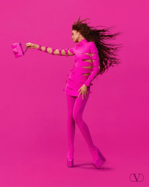 A woman in a pink dress and platform shoes holds a pink handbag against a pink background in a fashion advertisement.