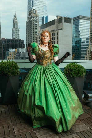 A woman in a voluminous green ball gown and sheer black gloves poses on a rooftop overlooking skyscrapers.