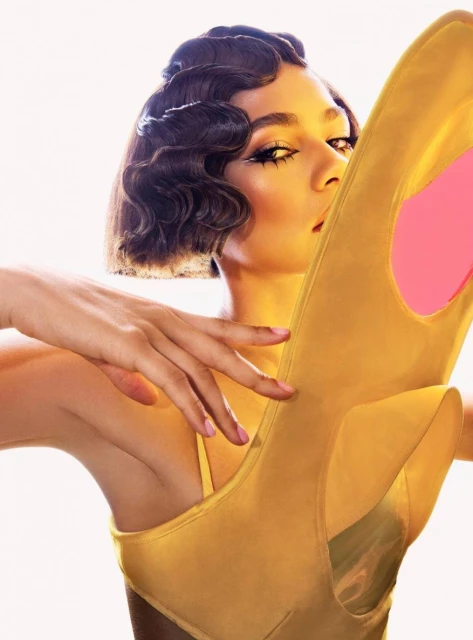 Close-up of woman with retro hairstyle and dramatic makeup, holding a yellow high-heel shoe in a glamour shot.