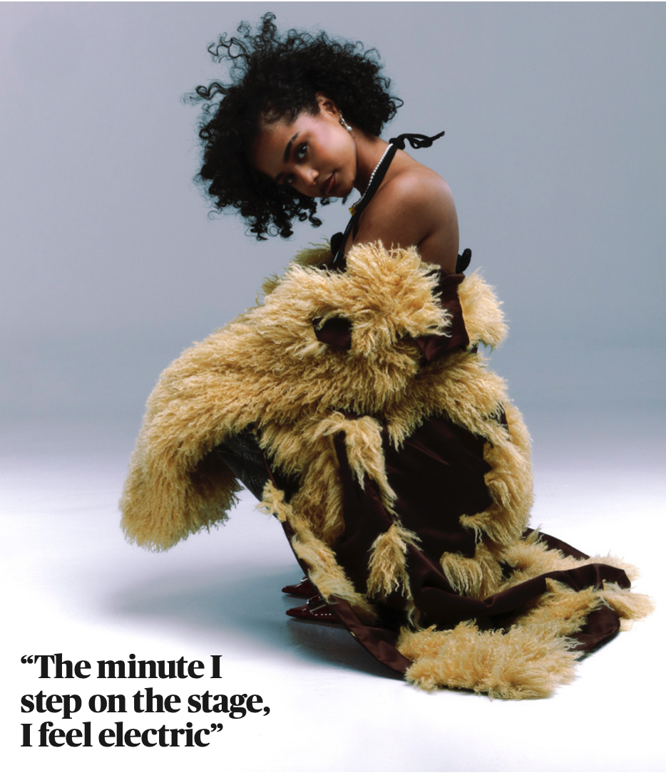 Model with dark curly hair wearing a brown garment with yellow faux-fur trim against a pale background